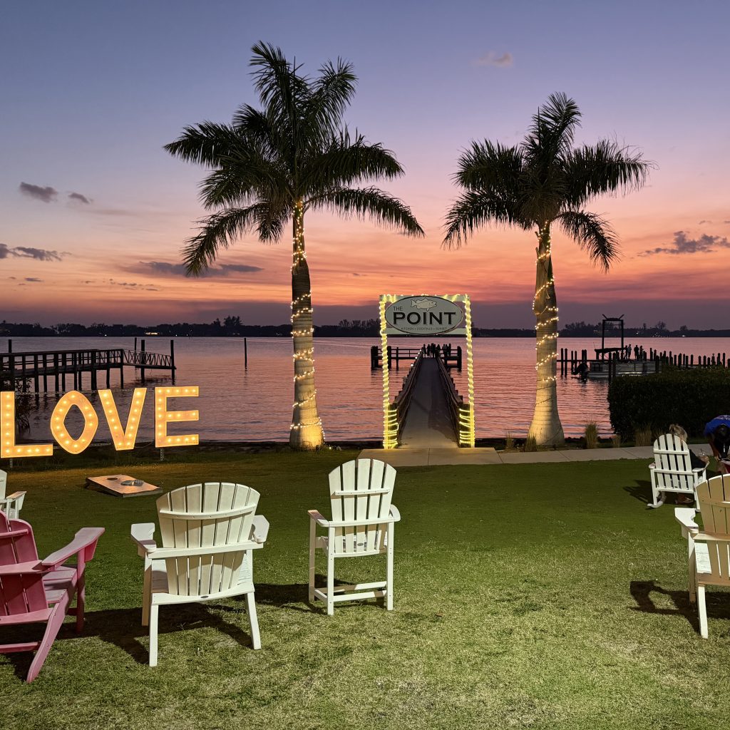 Evie’s On the Bay lawn with Adirondack chairs, palm trees, LOVE sign, dock, and sunset over the water.